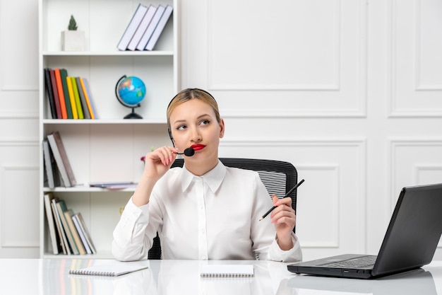 Service à la clientèle jolie fille blonde en chemise blanche avec ordinateur portable et casque regardant avec un stylo