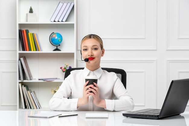 Service à la clientèle jolie fille blonde en chemise blanche avec ordinateur portable et casque tenant un gobelet en papier noir