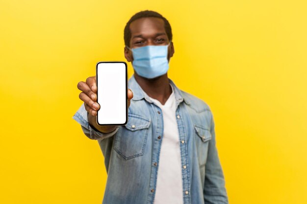 Service en ligne, technologie. Portrait d'un jeune homme satisfait et heureux avec un masque médical debout tenant un téléphone portable et souriant largement à la caméra. studio d'intérieur tourné isolé sur fond jaune