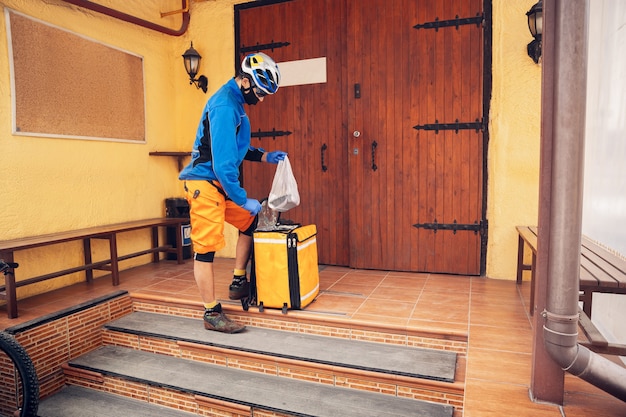 Service de livraison sans contact pendant la quarantaine. L'homme livre de la nourriture et des sacs à provisions pendant l'isolement. Frapper à la porte et laisser les marchandises jusqu'à ce que le client les récupère. Sécurité, réception, maintien à distance.