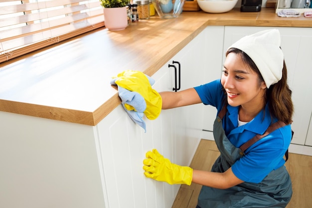 Service de nettoyage professionnel pour femme asiatique portant des gants en caoutchouc jaune à l'aide d'un chiffon pour essuyer avec un détergent liquide pulvérisé sur le comptoir de cuisine en bois à la maison