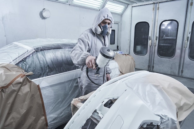 Service de peinture et de réparation automobile. Mécanicien automobile en salopette blanche peint voiture avec pulvérisateur aérographe dans la chambre de peinture