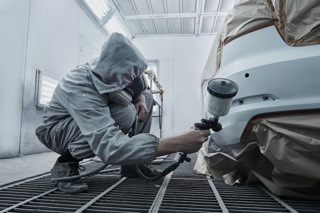 Service de peinture et de réparation automobile. Un mécanicien automobile en salopette blanche peint une voiture avec un pulvérisateur aérographe dans une chambre à peinture