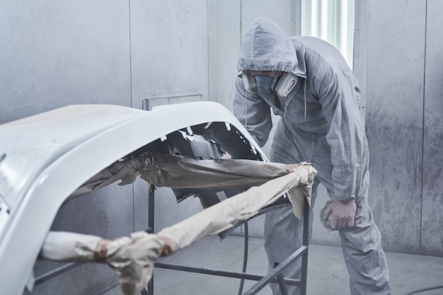 Service de peinture et de réparation automobile. Un mécanicien automobile en salopette blanche peint une voiture avec un pulvérisateur d'aérographe dans une chambre à peinture