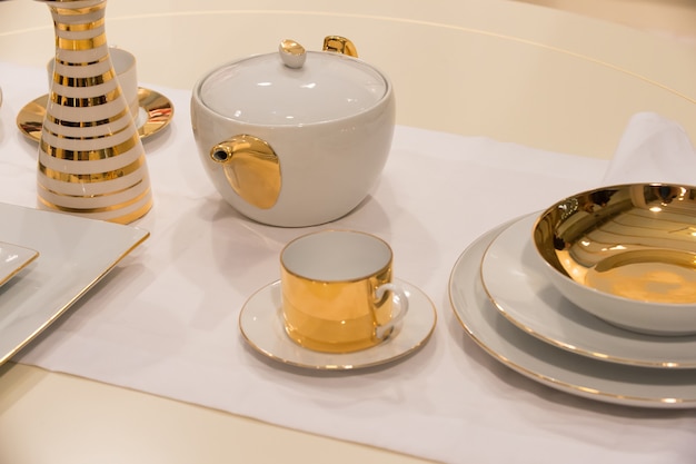 Service à thé ou ensemble avec pot, tasse, soucoupe, assiettes et vase avec dorure dorée servi sur fond de nappe blanche. Concept de thé ou de cérémonie