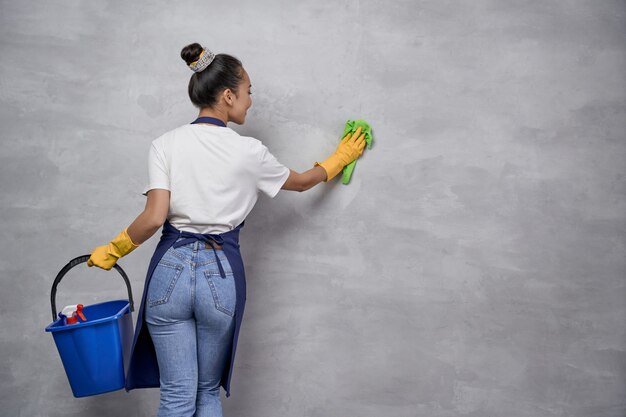 Services d'entretien ménager et de nettoyage. Vue arrière de l'uniforme de la femme au foyer ou de la femme de chambre et des gants en caoutchouc jaune tenant un seau ou un panier avec différents produits de nettoyage et nettoyant un mur. Désinfection