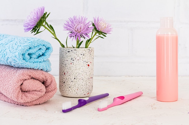 Des serviettes, des brosses à dents, une bouteille de shampoing et des fleurs d'asters sur une surface blanche