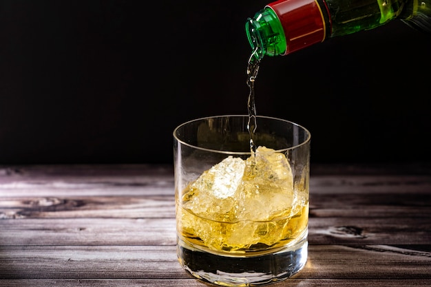 Servir du whisky à partir d'une bouteille dans un verre à whisky avec de gros morceaux de glace sur une table en bois rustique. Espace de copie.