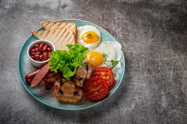 Servir un plat de la carte du restaurant. Petit-déjeuner délicieux et sain avec des œufs au plat, des haricots, des saucisses, des légumes grillés, des tomates et des champignons avec de la salade sur une assiette contre une table en pierre grise
