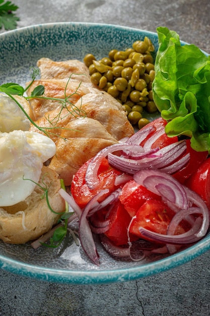 Servir un plat de la carte du restaurant. Poitrine de poulet aux légumes et légumes verts, tomates, oignons, omelettes et pois verts sur une assiette sur fond de table en pierre grise