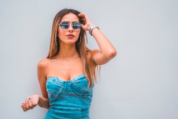 Session de style de vie, une jeune brunette caucasienne dans une robe en jean bleu et des lunettes de soleil sur un fond gris uni, avec place pour copier et coller