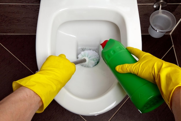 Un seul homme portant des gants en caoutchouc jaune nettoie la cuvette des toilettes avec une brosse et un désinfectant