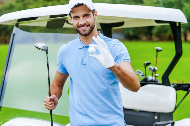 Seul le meilleur équipement de golf ! Beau jeune homme souriant tenant une balle de golf et un chauffeur tout en se tenant près de la voiturette de golf et sur le terrain de golf