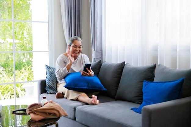 Seule une femme âgée asiatique à la retraite assise sur un canapé et parlant à sa famille par téléphone portable utilise Internet dans le salon pour se détendre à la maison