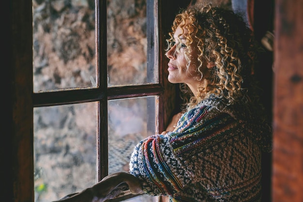 Une seule femme debout à la maison près de la fenêtre regardant à l'extérieur et pensant Mode de vie des femmes célibataires Attendre des amis et des pensées Jeune femme loisirs à l'intérieur Portrait latéral d'âge moyen