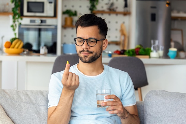 Une seule pilule peut aider Beau jeune homme tenant un verre d'eau et regardant une pilule dans sa main alors qu'il était assis sur le canapé à la maison