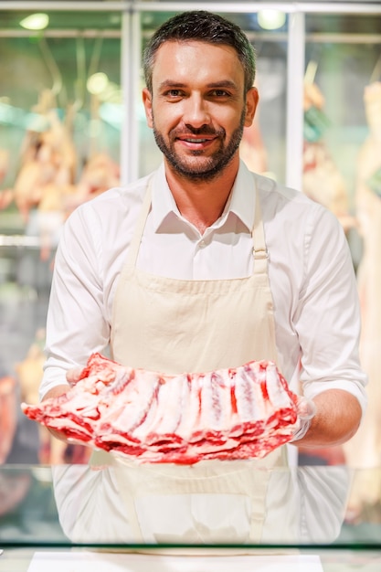 Seulement la viande la plus fraîche ! Souriant jeune boucher étirant une tranche de viande et regardant la caméra en se tenant debout à la caisse du supermarché