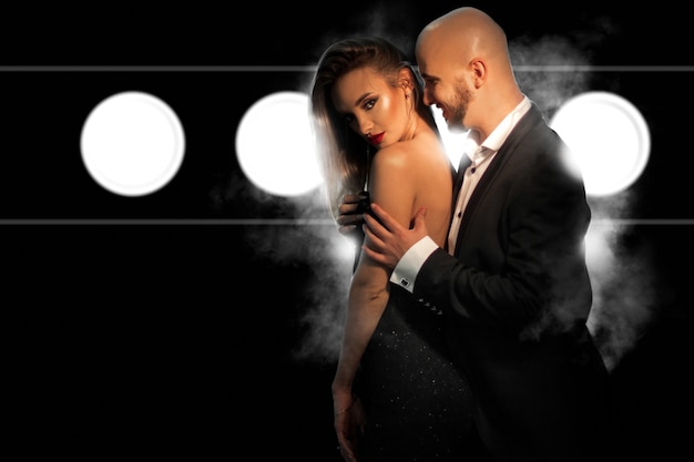 Sexuel jeune couple amoureux en costume noir et robe posant en studio sur un mur sombre avec de la fumée
