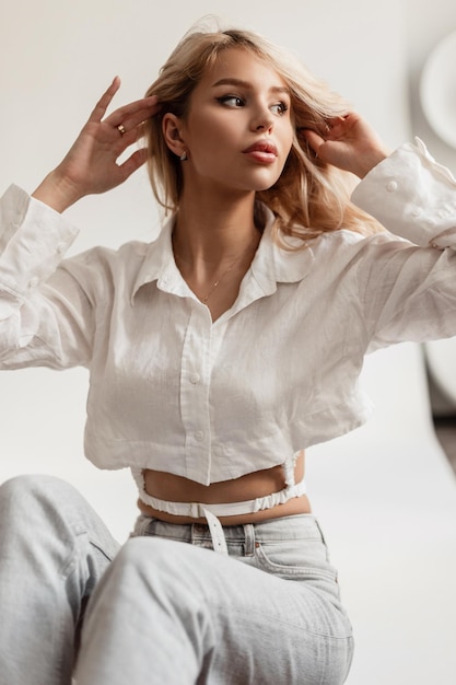 Sexy beau modèle aux cheveux blonds dans une chemise élégante et un jean est assis dans le studio et regarde de côté