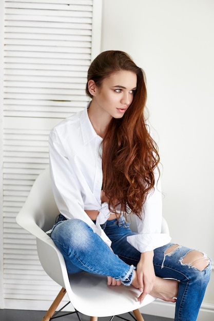 Sexy belle fille en chemise blanche jeans assis sur une chaise. Superbe jeune femme aux cheveux longs et aux yeux charmants. La femme d'affaires sexy est jeune et confiante. Fille assise sur une chaise à la maison, corps nu