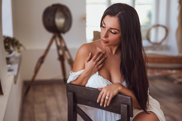 Sexy brunette assise dans une robe blanche près de la fenêtre