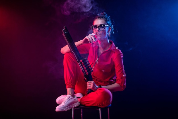 Sexy bully girl fume une cigarette avec un fusil de chasse sur un fond sombre à la lumière du néon et une brume de pâte de copie