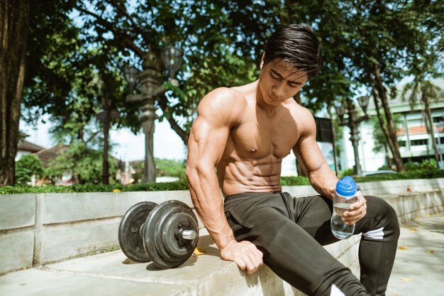 Sexy homme musclé assis pose tenant une bouteille d'eau près d'haltères tout en exerçant à l'extérieur dans le parc