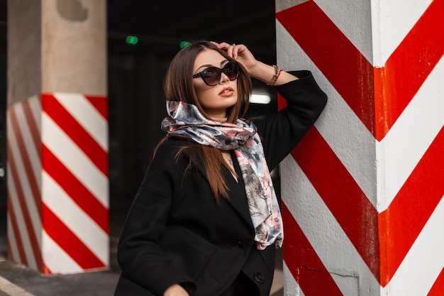 Sexy jeune femme brune à lunettes de soleil élégantes en manteau noir tendance avec foulard en soie ressort se repose près de la colonne rayée dans un parking de la ville. Mannequin élégante belle fille. Dame d'affaires moderne.