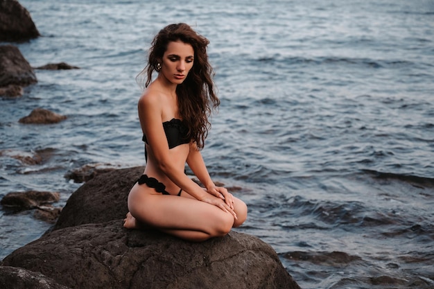 Sexy jeune fille en maillot de bain noir assis sur les rochers au bord de la mer en été