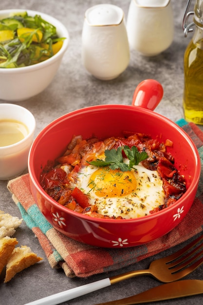 Shakshuka dans une casserole rouge, café, pain et salade dans un bol, fond gris, copy space