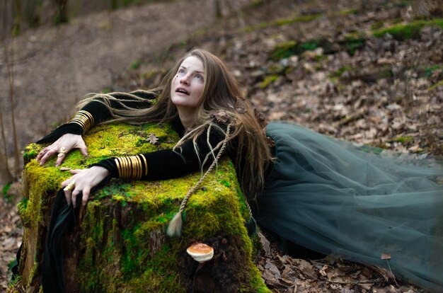 Shamane féminine dans la nature forestière vierge allongée sur une souche d'arbre verte bijoux en or cheveux poilues