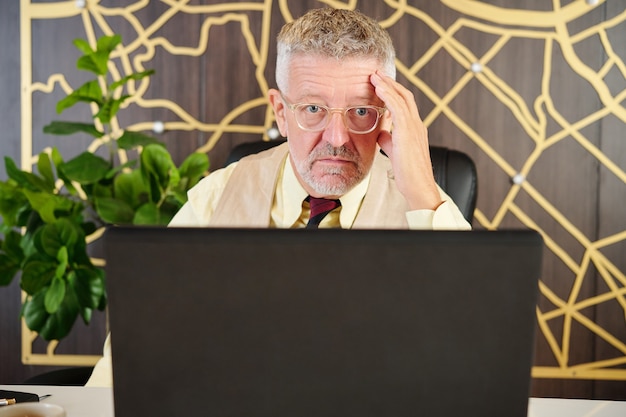 Shoked senior businessman reading report ou contrat sur écran d'ordinateur portable dans un bureau moderne