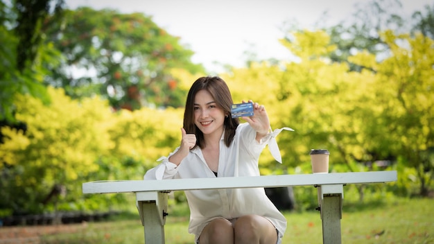 Shopper sourire fille achetant en ligne ou travaillant en ligne avec un téléphone intelligent et une carte de crédit assis sur un banc dans un parc de la ville en été Sourire fille heureuse avec sa carte portrait d'un jeune cadre