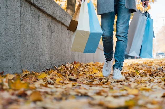 Shopping d'automne. Photo recadrée d'un homme marchant avec des colis le long d'un trottoir d'automne recouvert de feuilles jaunes. Copiez l'espace.