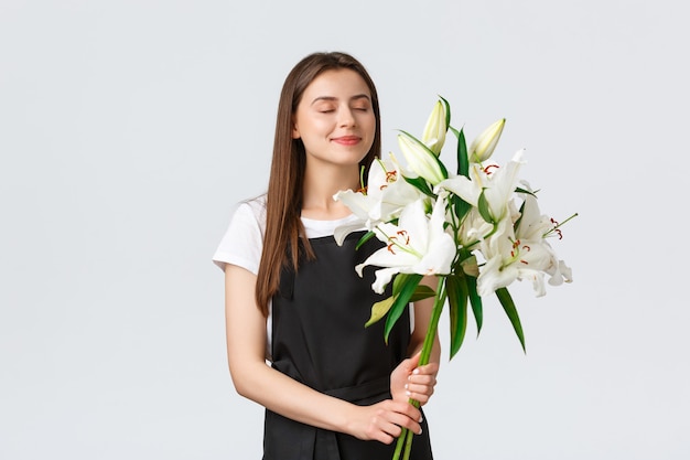 Shopping, employés et concept de petite entreprise. Une fleuriste souriante et rêveuse dans un magasin de fleurs ferme les yeux et renifle une bonne odeur de bouquet de lys blancs, fond blanc.