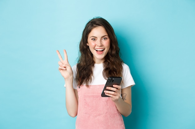 Shopping en ligne. Fille souriante positive montrant le signe v et l'air heureux devant la caméra, utilisant un smartphone, tenant un téléphone portable, debout sur fond bleu.