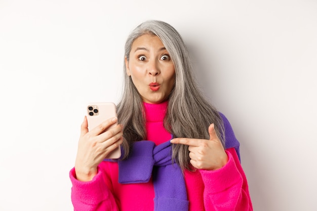 Shopping en ligne. Gros plan d'une femme âgée asiatique étonnée pointant sur un smartphone et souriante étonnée, debout sur fond blanc.