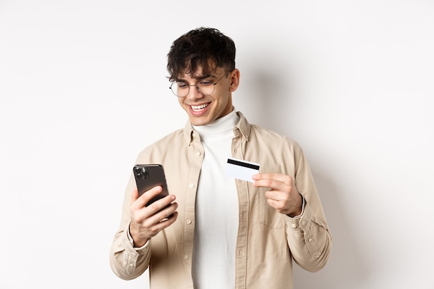 Shopping en ligne. Heureux jeune homme à l'aide de smartphone et de carte de crédit en plastique, payant sur internet, debout sur un mur blanc.