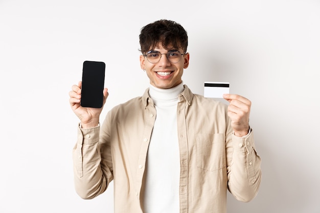 Shopping en ligne. Mec naturel dans des verres montrant l'écran du smartphone vide et une carte de crédit en plastique, souriant heureux, recommandant la banque, debout sur un mur blanc.