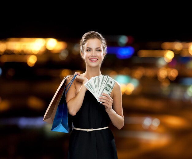 shopping, vente, cadeaux, argent et concept de vacances - femme souriante en robe avec des sacs à provisions et de l'argent sur fond noir