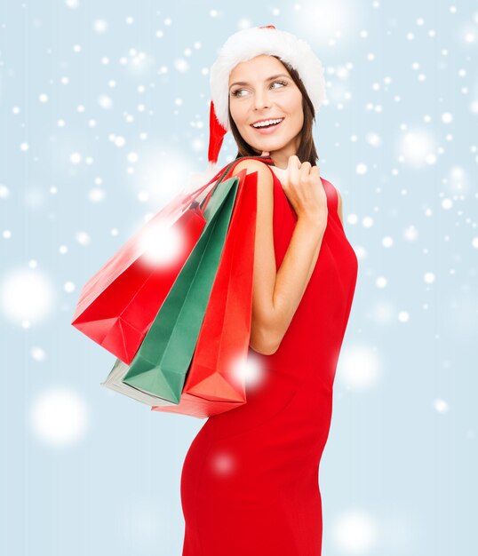 shopping, vente, cadeaux, noël, concept de noël - femme souriante en robe rouge et bonnet de noel avec des sacs à provisions