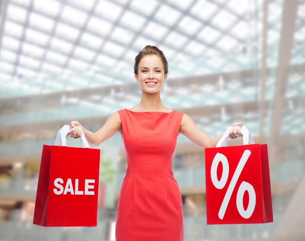 shopping, vente, cadeaux, noël et concept de vacances - jeune femme souriante en robe rouge avec des sacs à provisions avec signe de pourcentage et de vente