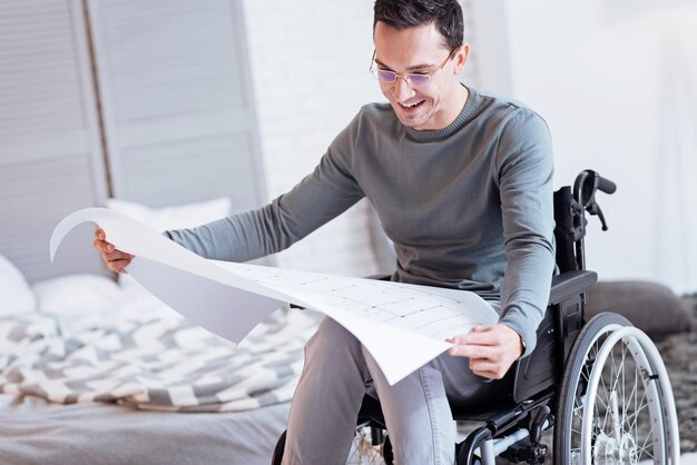 Si mignon. Homme invalide positif s'inclinant la tête et gardant le sourire sur son visage tout en étant en fauteuil roulant