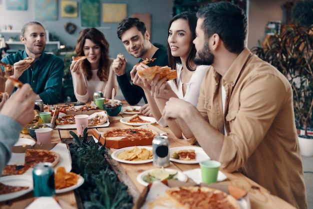 Si savoureux! Groupe de jeunes en tenue décontractée mangeant de la pizza et souriant tout en dînant à l'intérieur