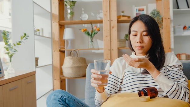 Sick Asian young woman holding pill prendre un médicament à la recherche assis sur le canapé à la maison