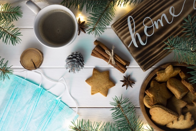 Signe de bois de lettrage à la maison avec biscuit de pain d'épice, branches d'arbre de Noël, tasse de café, bâtons de cannelle et masques médicaux sur une surface blanche. Mise à plat.