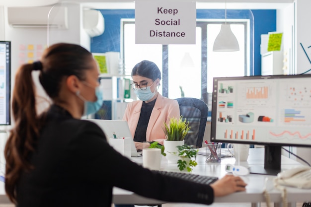 Signez avec garder la distance sociale dans le nouveau bureau normal pendant la pandémie mondiale entre collègues, portant un masque facial comme prévention de la sécurité. Femme analysant les statistiques financières en milieu de travail.