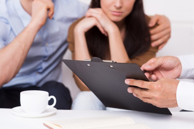 Signez ici. Gros plan d'un jeune couple réfléchi assis sur le canapé tandis que l'homme en tenue de soirée pointant le presse-papiers avec le document