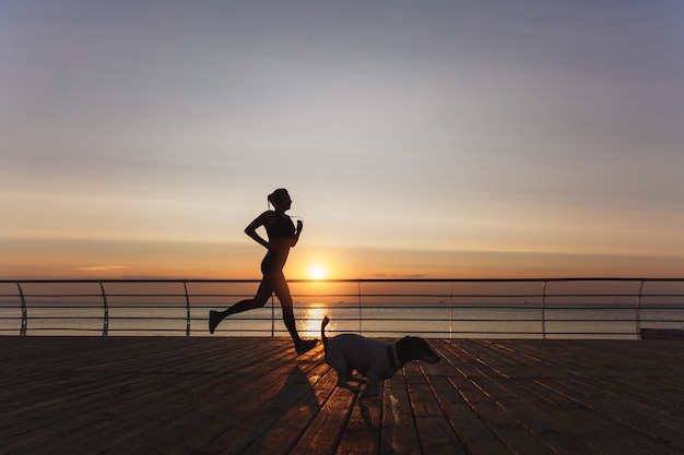 La silhouette d'une belle jeune fille athlétique aux longs cheveux blonds dans un casque qui court à l'aube sur la mer avec son chien