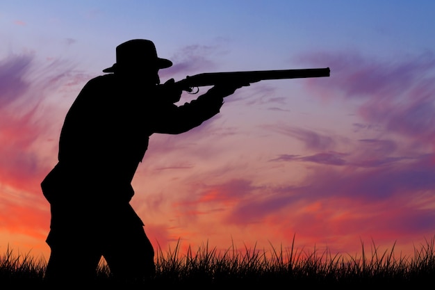 Silhouette d'un chasseur avec une arme à feu dans les roseaux contre le soleil, une embuscade pour les canards avec des chiens